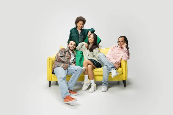 Happy multiracial friends in trendy outfit posing on yellow couch on grey background — Stock Photo