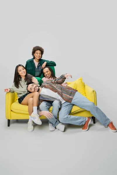 Радісні мультикультурні друзі в модному вбранні розважаються на жовтому дивані на сірому фоні — стокове фото