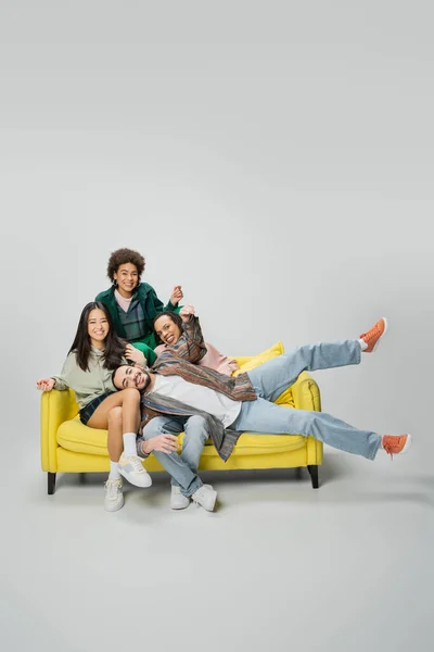 Веселые многонациональные друзья, смотрящие в камеру и веселящиеся на желтом диване на сером фоне — стоковое фото