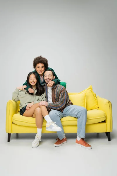 Cheerful african american woman embracing stylish multiethnic friends sitting on yellow couch on grey background — Stockfoto