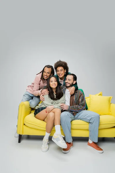 Sorridenti amici multirazziali in abiti alla moda in posa vicino divano giallo su sfondo grigio — Foto stock