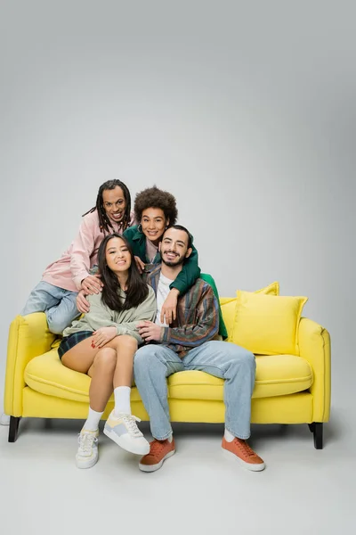 Happy and stylish multicultural friends posing near yellow couch and looking at camera on grey background — Stock Photo
