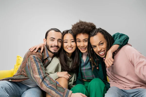 Felice donna afro-americana che abbraccia felici amici multietnici seduti sul divano e sorridenti alla telecamera isolata sul grigio — Foto stock