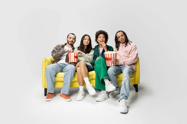 Amazed african american woman eating popcorn while watching movie with multiethnic friends on grey background — Stock Photo