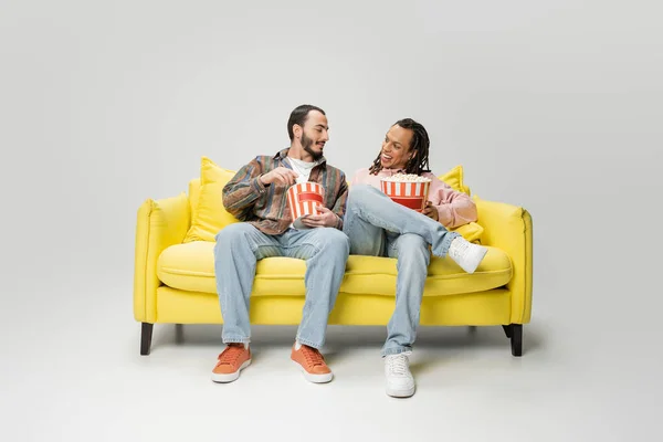 Full length of stylish multiethnic men smiling at each other while sitting with buckets of popcorn on yellow couch on grey background — Stock Photo