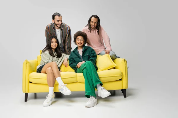 Cheerful african american women sitting on yellow couch near trendy multiethnic men on grey background — Stock Photo