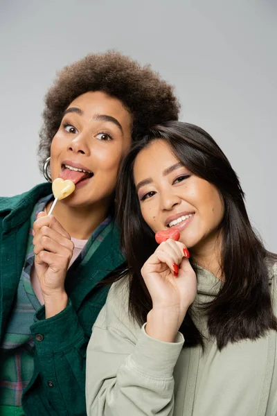 Cheerful african american woman with lollipop sticking out tongue near asian friend isolated on grey — Stockfoto