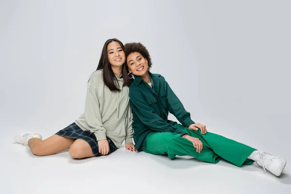 Full length of asian and african american women in trendy outfit sitting and looking at camera on grey background — Stock Photo