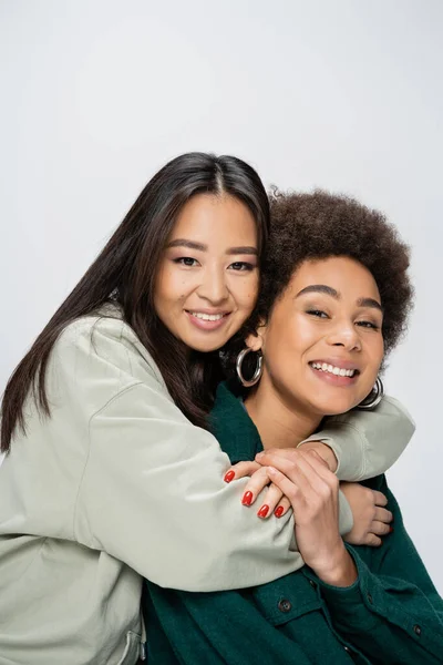 Happy asian woman embracing curly african american friend while looking at camera isolated on grey — Stock Photo