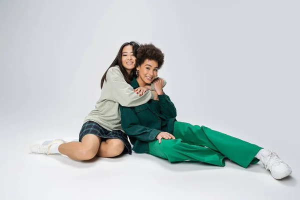 Full length of happy asian woman embracing stylish african american friend while sitting on grey background — Stock Photo