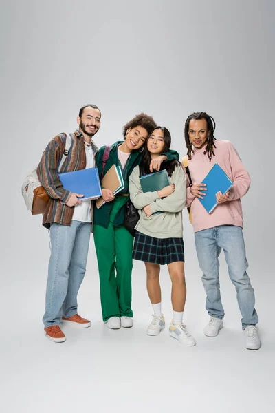 Full length of happy multiethnic students with backpacks and notebooks looking at camera on grey background — Stock Photo