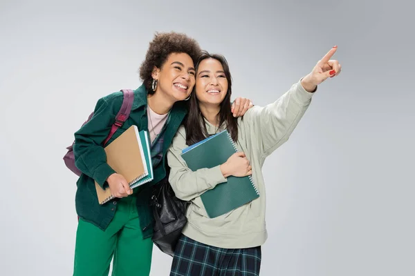 Smiling asian student with copybooks looking away and pointing with finger near cheerful african american friend isolated on grey — Stock Photo
