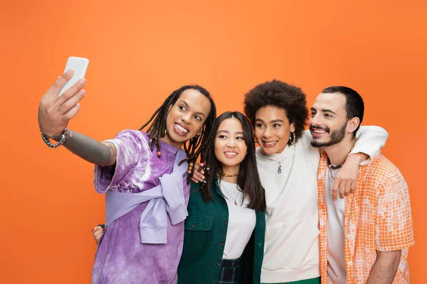 Uomo alla moda con dreadlocks scattare selfie con amici multiculturali felici isolati su arancione — Foto stock