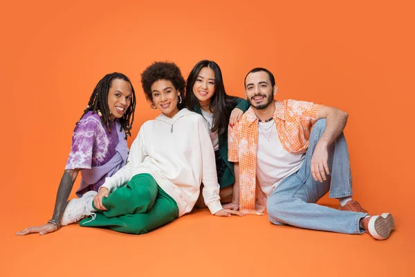 Joyful multicultural friends in stylish clothes sitting and looking at camera on orange background — Stockfoto