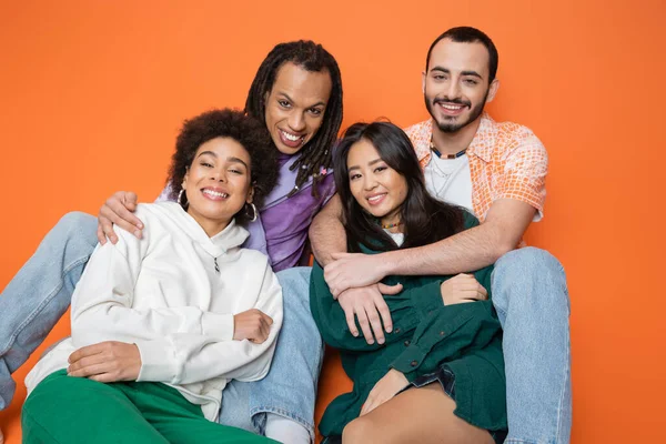Amigos multiculturais positivos em roupas da moda sorrindo para a câmera enquanto sentado no fundo laranja — Fotografia de Stock
