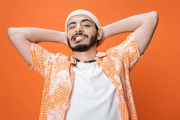 Stylish bearded man in white beanie holding hands behind head and grimacing isolated on orange — Stock Photo
