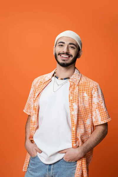 Young bearded man in white beanie and beads posing with hands in pockets of jeans isolated on orange — Stockfoto