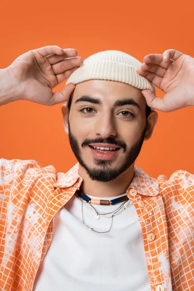 Portrait of young bearded man adjusting white beanie and smiling at camera isolated on orange — Stock Photo