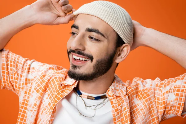 Portrait of cheerful bearded man in beads touching white beanie and looking away isolated on orange — Stock Photo