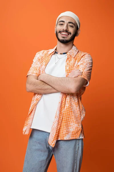 Happy bearded man in trendy shirt and white beanie posing with crossed arms isolated on orange — Stock Photo