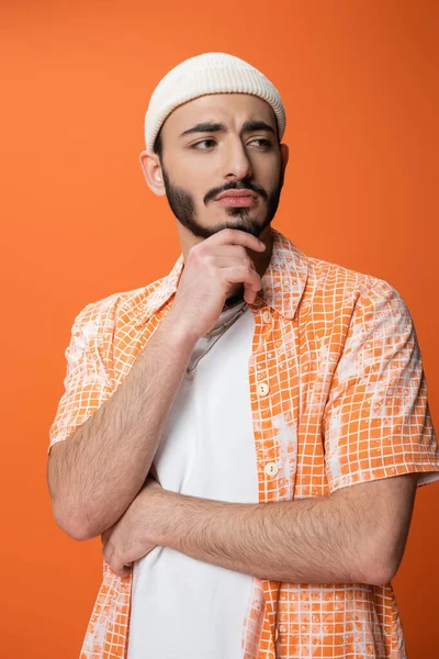 Upset bearded man in stylish shirt and white beanie holding hand near chin and looking away isolated on orange — Stock Photo