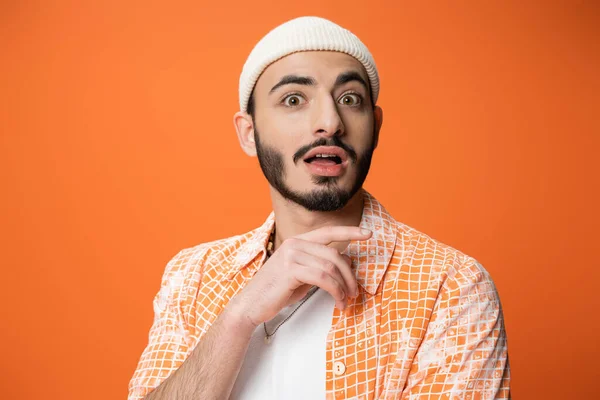 Impressed bearded man in trendy shirt and white beanie looking at camera isolated on orange — Stock Photo