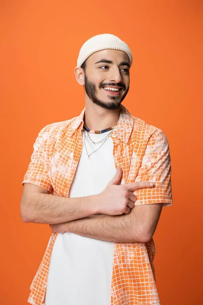 Homme barbu à la mode souriant et pointant du doigt tout en regardant loin isolé sur orange — Photo de stock