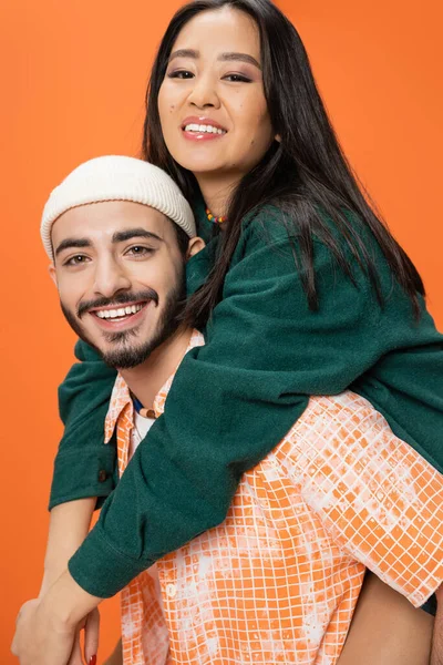 Heureux asiatique femme et jeune barbu homme dans bonnet sourire à caméra tout en s'amusant isolé sur orange — Photo de stock