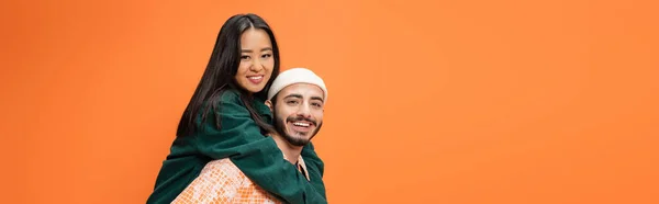 Feliz barbudo hombre en beanie piggybacking bastante asiático mujer aislado en naranja, bandera — Stock Photo