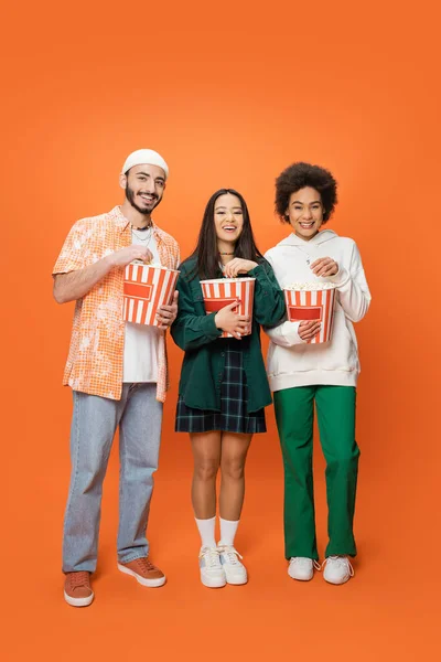 Pleine longueur d'amis multiethniques branchés debout avec des seaux de maïs soufflé et souriant à la caméra sur fond orange — Photo de stock