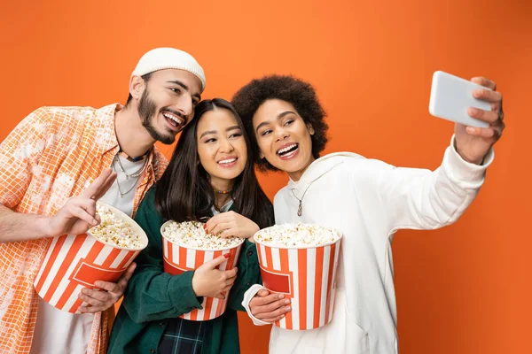 Happy african american woman taking selfie with trendy friends holding buckets of popcorn isolated on orange — Stock Photo