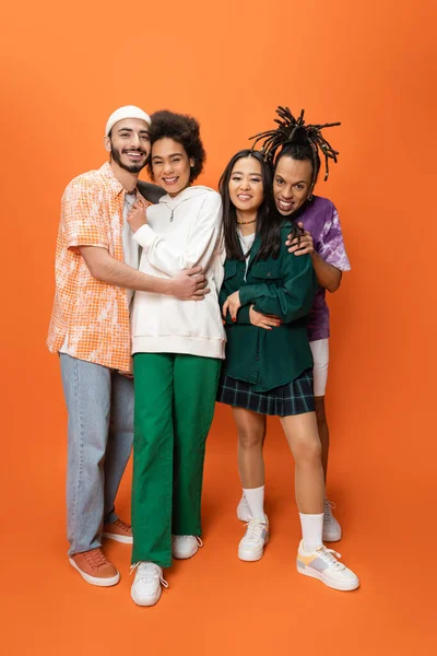 Volle Länge von modischen multikulturellen Freunden, die sich umarmen und in die Kamera auf orangefarbenem Hintergrund schauen — Stockfoto