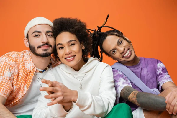 Amigos multiétnicos felizes na roupa na moda sentado e olhando para a câmera isolada em laranja — Fotografia de Stock