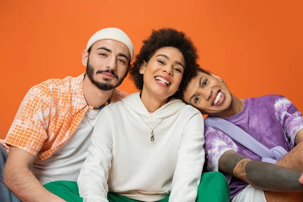 Amigos multiculturais felizes em traje elegante olhando para a câmera enquanto sentado isolado em laranja — Fotografia de Stock