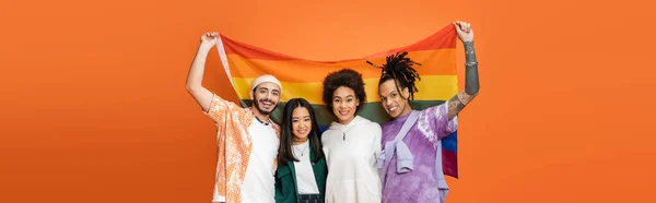 Amigos multiétnicos felizes em roupas da moda segurando bandeira lgbt isolado em laranja, bandeira — Fotografia de Stock