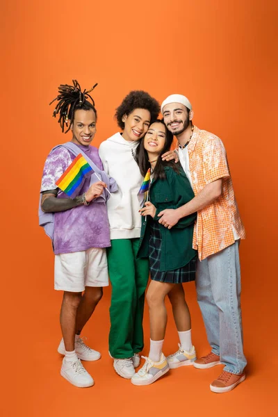 Full length of stylish and cheerful multicultural friends embracing while holding lgbt flags on orange background — Stock Photo