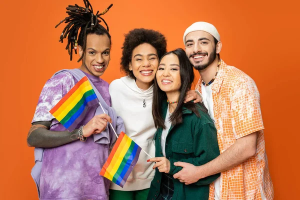Multicultural friends in stylish clothes holding lgbt flags and smiling at camera isolated on orange — Stock Photo