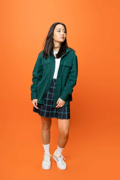 Full length of young asian woman in checkered skirt and green shirt looking away on orange background — Stockfoto