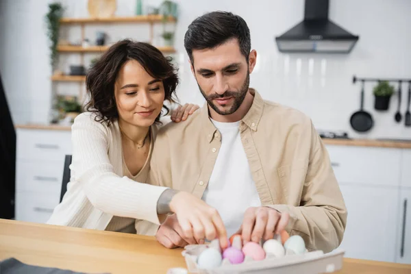 Smiling woman and man putting Easter eggs in tray at home — Fotografia de Stock