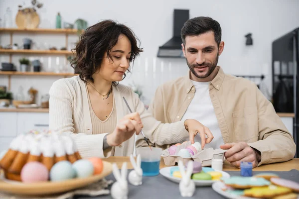 Woman holding paintbrush and taking Easter egg near husband in kitchen — Fotografia de Stock