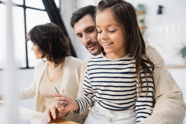 Smiling girl holding paintbrush near blurred parents at home — Fotografia de Stock