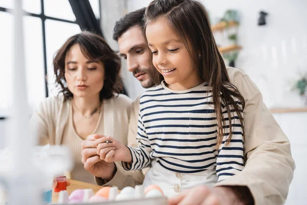 Smiling girl holding paintbrush near blurred Easter eggs and parents at home — Stock Photo