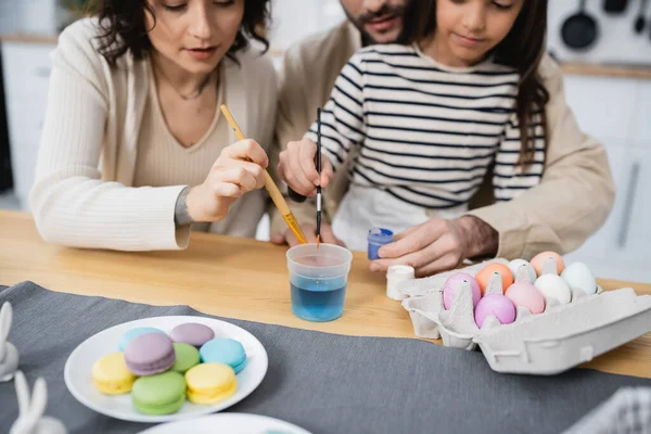 Vista recortada de padres borrosos y niños sosteniendo pinceles cerca de macarrones y huevos de Pascua en casa - foto de stock