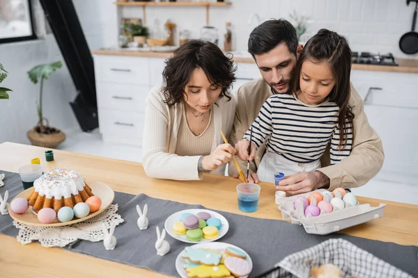 Família colorir ovos de Páscoa perto de bolo e biscoitos na cozinha — Fotografia de Stock