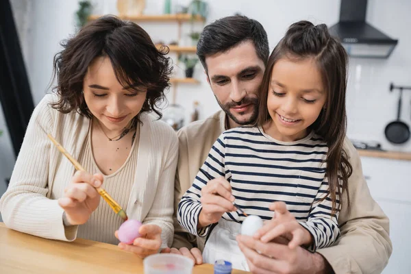 Familia feliz sonriendo mientras colorea huevos Easer en la cocina - foto de stock