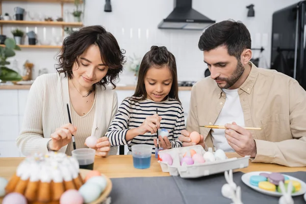 Família pintura ovos de Páscoa perto de bolo borrado na mesa na cozinha — Fotografia de Stock