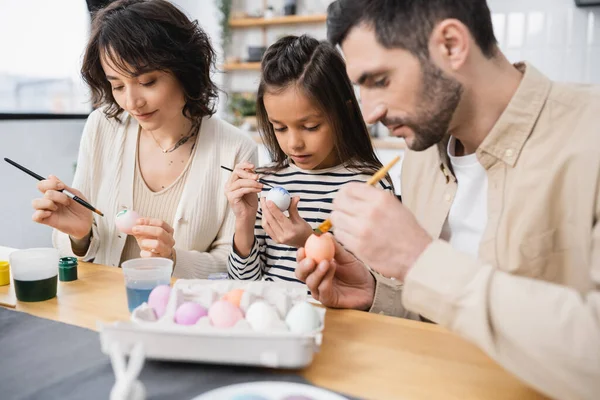 Concentrated girl painting Easter egg near parents in kitchen — Stock Photo