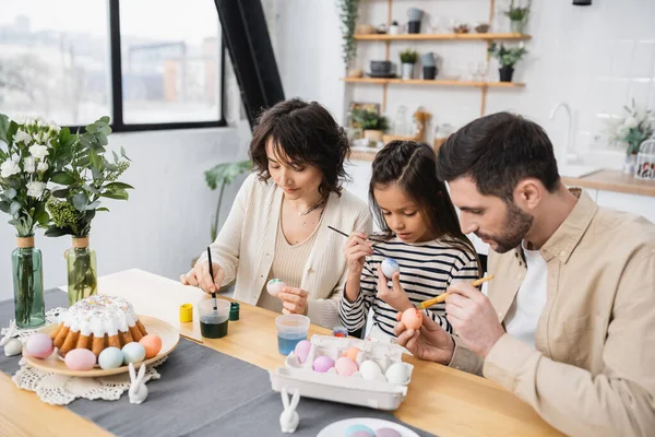 Familia para colorear huevos de Pascua cerca de sabrosa torta en la mesa en casa - foto de stock