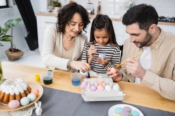 Усміхнена жінка розмальовує великодні яйця біля сім'ї та їжі вдома — стокове фото