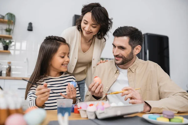 Pais felizes olhando para a filha colorir ovo de Páscoa em casa — Fotografia de Stock